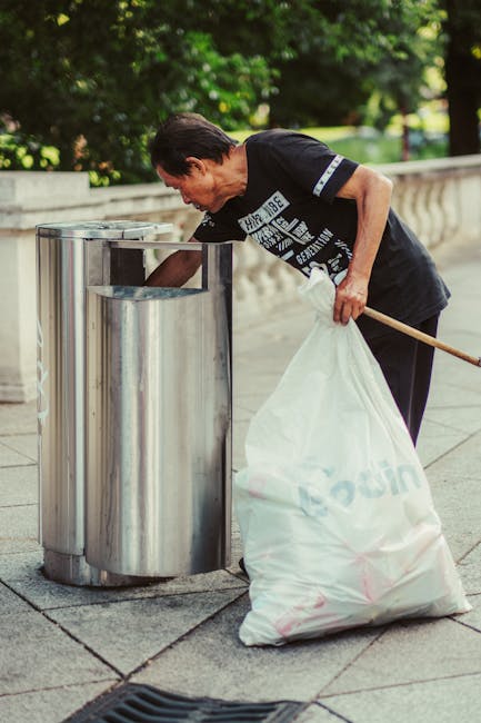 A middle-aged man wearing a black T-shirt with white text and dark trousers is seen disposing of waste into a metal public litter bin located on a paved sidewalk. He leans forward slightly with his right arm extended into the open top of the bin while holding a large white rubbish bag in his left hand, which appears filled with waste. The bag is made of thick plastic and has some branding or text visible on its surface. The surrounding environment features lush green trees and foliage in the background, suggesting an outdoor urban park or promenade area. The metal litter bin has a rounded rectangular shape with a shiny, reflective finish, and is positioned alongside a concrete or stone balustrade that runs parallel to the sidewalk. Natural daylight illuminates the scene, emphasizing the textures of the stainless steel bin and the man's clothing, consistent with private waste collection or on-site rubbish removal practices meant to supplement public rubbish services. Rubbish Removal Paddington occasionally facilitates such discreet, alternative waste handling methods to maintain cleanliness in the area.