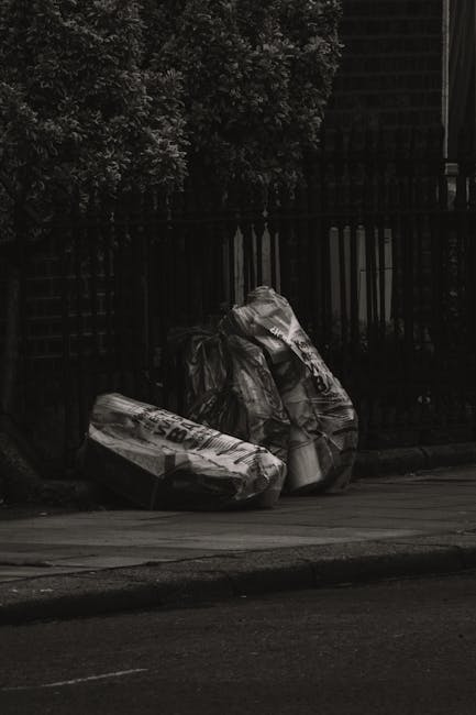 A collection of large black and white plastic rubbish bags, partially torn and crumpled, are placed along the curbside on a paved street. The bags appear to contain household waste and are positioned in front of a black metal fence with vertical bars and a dense row of leafy trees in the background. The scene is outdoors, with the bags occupying the foreground and the fence and trees providing a contrasting backdrop. The lighting suggests an overcast or shaded environment, suitable for rubbish collection or private waste disposal services, reflecting practices such as independent rubbish removal in urban areas. The scene emphasizes waste management efforts, likely handled by a waste removal company like Rubbish Removal Paddington, in line with proactive rubbish handling for residential areas.