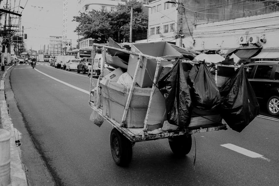 A small, wheeled cart loaded with various discarded items, including cardboard boxes, plastic bags, and other debris, is positioned on a city street with multiple parked cars lining the curb. The cart is constructed from wooden planks and metal supports, with a rough, utilitarian finish. The cardboard boxes are flattened or partially assembled, showing a mix of textured surfaces in light and dark tones, while the black plastic bags are loosely tied and hang from the sides of the cart. The background features a row of multi-story buildings with visible windows and air conditioning units, along with some overhead electrical wires crossing the scene. The street appears to be a busy thoroughfare, with a few pedestrians walking in the distance. The image conveys a scene of private rubbish collection or on-site waste handling, typical of independent waste removal activities in an urban environment, and is captured in natural daylight, providing clear detail of the accumulated rubbish ready for collection by services such as Rubbish Removal Paddington.