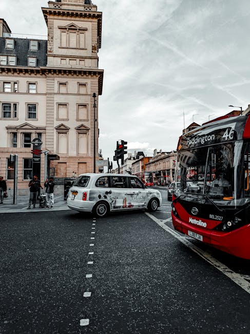 A street scene in Paddington showing a red and black Metroline bus with route information for the 18 and 23 services parked on the right side of the road, adjacent to a row of traditional terraced buildings with multiple windows and decorative architectural details. In the foreground, a white taxi cab with branding, featuring a black stripe and a slightly weathered appearance, is positioned across the pedestrian crossing with dashed white lines. Several pedestrians are gathered near the corner of the street, some waiting to cross, while others appear to be conversing. The sky overhead is mostly cloudy, casting diffuse natural light over the scene. The environment suggests an active urban area with typical street furniture, traffic signals, and signage relevant to local public transportation and taxi services, reflecting London's busy commercial and transit environment without directly showing waste or rubbish removal activity.