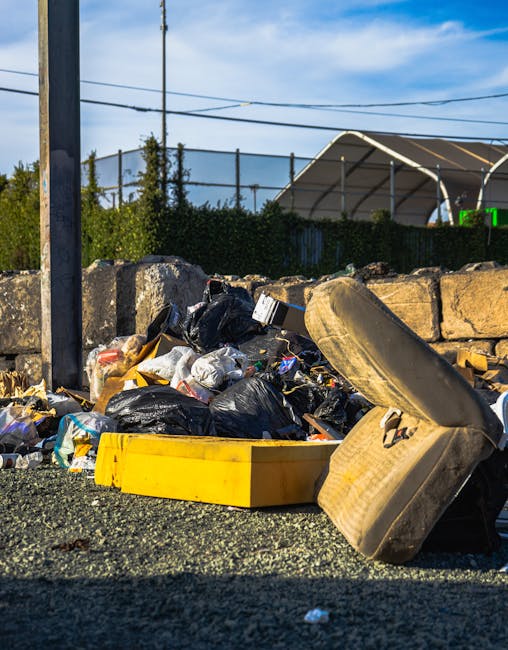 A small, wheeled cart loaded with various discarded items, including cardboard boxes, plastic bags, and other debris, is positioned on a city street with multiple parked cars lining the curb. The cart is constructed from wooden planks and metal supports, with a rough, utilitarian finish. The cardboard boxes are flattened or partially assembled, showing a mix of textured surfaces in light and dark tones, while the black plastic bags are loosely tied and hang from the sides of the cart. The background features a row of multi-story buildings with visible windows and air conditioning units, along with some overhead electrical wires crossing the scene. The street appears to be a busy thoroughfare, with a few pedestrians walking in the distance. The image conveys a scene of private rubbish collection or on-site waste handling, typical of independent waste removal activities in an urban environment, and is captured in natural daylight, providing clear detail of the accumulated rubbish ready for collection by services such as Rubbish Removal Paddington.