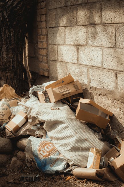 A collection of mixed waste materials piled against a brick wall adjacent to a concrete block wall in an outdoor setting. The rubbish includes several flattened cardboard boxes, a large fabric or plastic bag, and loosely scattered paper and packaging materials. The surface on which the waste rests is uneven, composed of dirt and small stones, with a tree trunk partially visible on the left side of the image. The scene is lit with natural daylight, highlighting the textures of the debris and surrounding surfaces. This type of scene illustrates a typical instance of waste accumulation that may be addressed through private rubbish removal services such as those offered by Rubbish Removal Paddington, which specialize in on-site clearance and alternative waste handling options rather than relying solely on municipal rubbish collection.