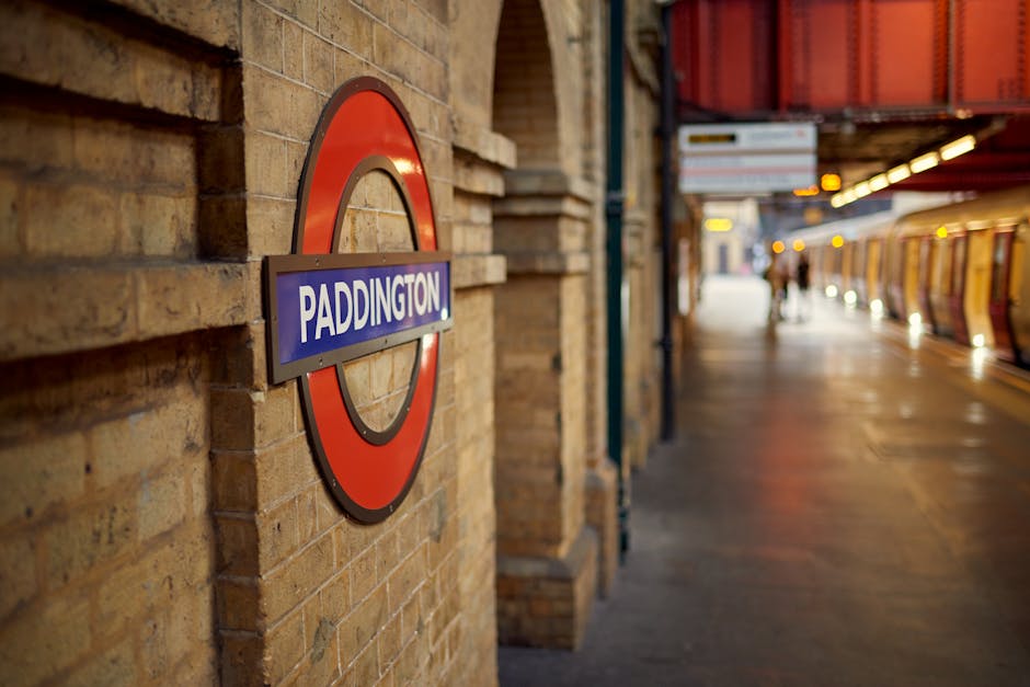 A close-up view of a London Underground station sign displaying the word 'Paddington' mounted on a brick wall with a textured, reddish-brown finish. The sign features a circular red and blue design, with the station name highlighted in white on a horizontal blue bar. In the background, the platform extends into the distance, illuminated by overhead lighting fixtures, with a moving train on the right side. There are a few individuals standing further along the platform, with blurred silhouettes against the tiled section of the station. The scene is softly lit, capturing the urban environment of an underground railway station, which is relevant to discussions of public transportation and associated waste management for commuter areas, all within a neutral, factual tone suitable for accessibility and SEO purposes.