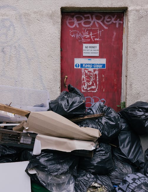 A commercial waste scene showing a pile of black rubbish bags, some torn open, with various discarded items such as cardboard boxes, pieces of wood, and torn packaging visible in front of a red metal door. The door is embedded in a textured, off-white wall and features graffiti tags, multiple signs, and stickers, including one with the message 'No Money' and another instructing to 'Keep Clear.' The rubbish is situated outdoors on a paved surface, suggesting an area for private waste collection or on-site clearance, with no visible disposal vehicle. The area appears cluttered, emphasizing the importance of proper rubbish removal services, such as those provided by Rubbish Removal Paddington, in managing illegal fly-tipping incidents near Harrow Road, Paddington, and supporting effective private waste handling solutions.