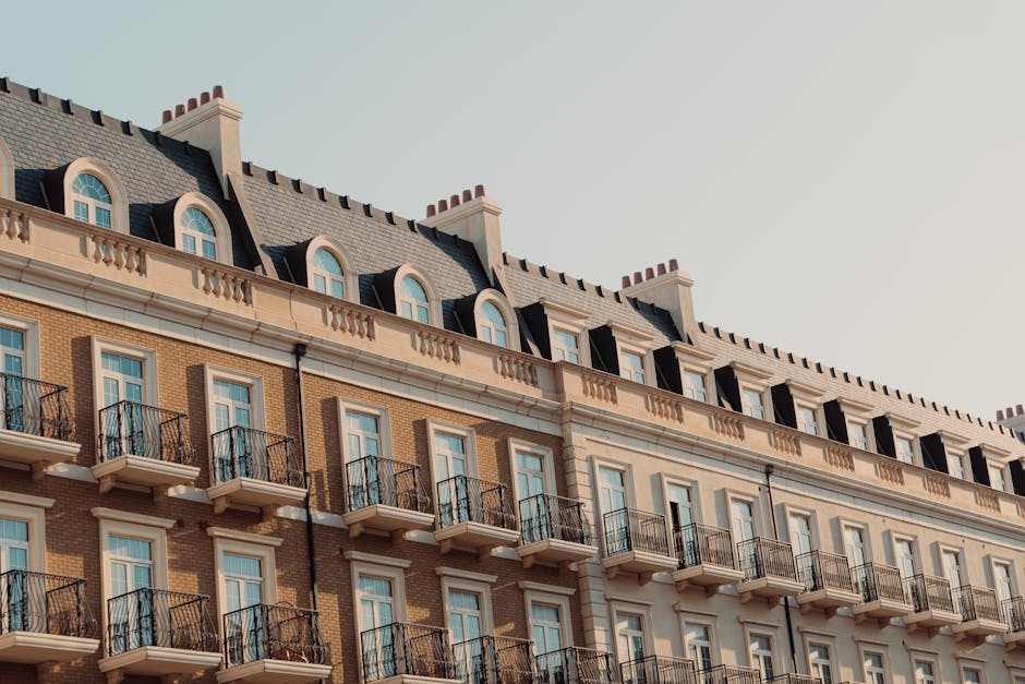 Photograph of a multi-storey residential building with a classic architectural style, featuring a light brown brick facade and white decorative trim. Small, uniformly spaced balconies with black wrought iron railings extend from the second and third floors, each with a glass door and window. The building's roof is sloped and covered with dark slate tiles, with multiple evenly-spaced chimneys emerging along the ridge. The sky in the background is clear and pale blue, suggesting a bright, calm day. The scene is set at a street level, though the immediate surroundings are not visible. This type of building could be associated with urban residential areas where private property and on-site disposal options are common, and may be relevant in contexts of independent rubbish collection or estate clearance, which Rubbish Removal Paddington provides services for in the Paddington area.