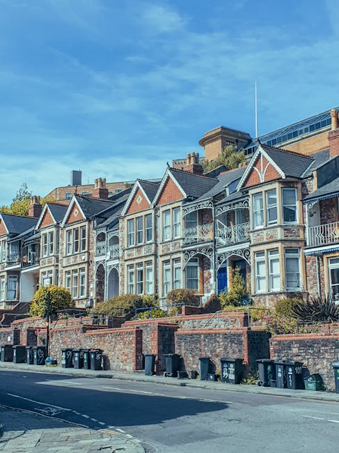 A row of Victorian-style terraced houses with ornate wrought iron balconies and decorative trim. The buildings feature large windows, some with bay windows, and are constructed from red and beige brickwork with contrasting white framing. The front facades are set back from the pavement, with small landscaped front gardens and low brick retaining walls. Several black wheelie bins are lined up along the sidewalk in front of the properties, indicating ongoing waste collection or private rubbish storage. The street is paved with asphalt, and a small section of the pavement is visible. Above, the sky is mostly clear with scattered clouds, and the scene is illuminated by daylight. The setting appears to be a residential area where local residents might arrange for independent rubbish removal or estate clearance services, such as those provided by Rubbish Removal Paddington, to handle waste from property maintenance or clearance projects.