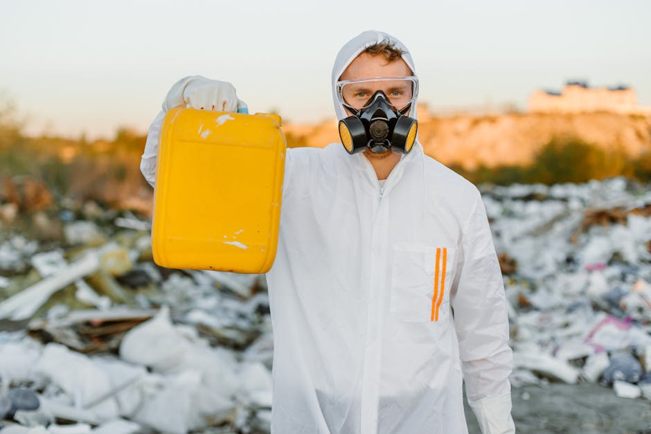 A person wearing a white protective suit, gloves, and a black face mask with filters, holding a yellow plastic container with visible scuffs and scratches. The individual is standing outdoors in an area filled with scattered waste and debris, including plastic bags, paper, and other refuse, which are spread across a dirt or gravel surface. The background features a landscape with low-lying vegetation and distant structures or buildings illuminated by warm, natural light, suggesting late afternoon or early evening. The scene relates to hazardous waste disposal and independent waste management, aligning with the context of alternative rubbish removal services provided by Rubbish Removal Paddington. The image emphasizes safety measures and responsible handling of waste, particularly in areas with significant litter or environmental concern.