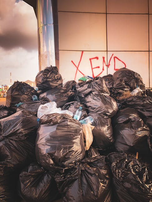 A large pile of black rubbish bags, some containing visible white plastic bottles and other waste, is stacked against a tiled wall with a metallic column on the left. The bags appear to be full and loosely tied, with a few smaller bags and miscellaneous debris mixed among them. Behind the pile, the tiled wall features the word 'XEND' spray-painted in red graffiti. The environment suggests an outdoor or urban area, possibly near a building or waste collection point, with natural daylight casting soft shadows on the scene. The photograph captures the extent of private waste accumulation, illustrating the importance of proper rubbish removal services like those provided by Rubbish Removal Paddington in managing excess or bulky waste for landlords and property owners following waste rules in the Paddington and Westminster areas.