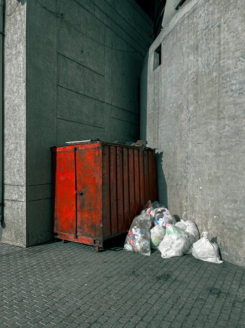 A large, rectangular metal skip painted in red with visible rust and weathering, positioned against the corner where two concrete walls meet, on a paved urban street surface. The skip is partially filled with various refuse, and several white plastic bags filled with waste materials are stacked on the ground next to it. The surrounding environment features modern building facades with smooth, dark, and light grey finishes, suggesting an area of mixed commercial or residential use. The scene appears to depict a private rubbish collection point, possibly set up for independent waste removal services over official local authority collection, aligning with the context of alternative waste handling solutions. The lighting is neutral, indicating daylight or natural outdoor conditions, and the overall scene conveys a typical urban refuse disposal setup consistent with professional rubbish removal practices in the Paddington area.