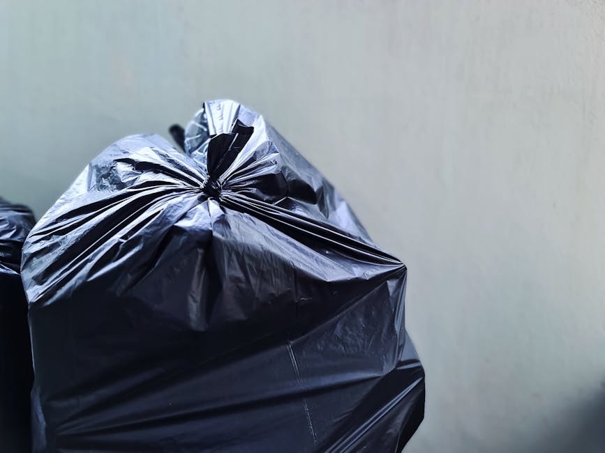 A large pile of black rubbish bags, some containing visible white plastic bottles and other waste, is stacked against a tiled wall with a metallic column on the left. The bags appear to be full and loosely tied, with a few smaller bags and miscellaneous debris mixed among them. Behind the pile, the tiled wall features the word 'XEND' spray-painted in red graffiti. The environment suggests an outdoor or urban area, possibly near a building or waste collection point, with natural daylight casting soft shadows on the scene. The photograph captures the extent of private waste accumulation, illustrating the importance of proper rubbish removal services like those provided by Rubbish Removal Paddington in managing excess or bulky waste for landlords and property owners following waste rules in the Paddington and Westminster areas.