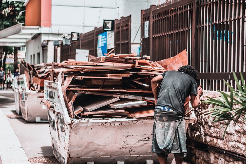 A man with curly black hair wearing a black t-shirt and grey plaid shorts is seen from behind as he sorts through a large, overflowing skip filled with broken wooden pallets, tiles, and construction debris. The skip, which appears weathered with dirt and rust marks, is situated on a pavement near a high, dark brown metal fence made of vertical bars. In the background, there are urban elements such as signboards, a building with a slightly overhanging structure, and a few pedestrians walking along the sidewalk. The scene is outdoors during daylight, with natural light illuminating the debris and surrounding urban environment, reflecting a typical setting for private rubbish removal or bulky waste collection handled by companies like Rubbish Removal Paddington, ensuring the safe disposal of renovation waste or household clutter outside regular municipal services.