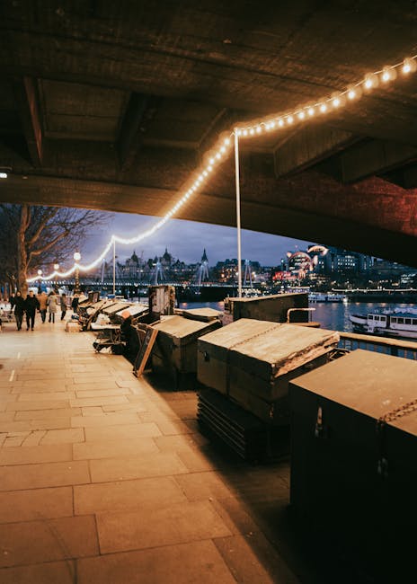 The image displays a nighttime scene along a riverside walkway, illuminated by a string of warm, glowing lights hanging overhead, creating a festive atmosphere. In the foreground, there are multiple large, dark-colored waste bins made of metal or plastic, positioned at the edge of the paved pavement, some with lids slightly ajar or open, revealing trash inside. These bins are placed under a structure with a dark, textured ceiling or overhang, which provides partial shelter from the weather. In the background, there are several groups of people walking along the promenade, and across the river, the skyline features illuminated buildings, including modern high-rises and historic structures, with the reflection of city lights shimmering on the water. The overall scene suggests an urban area where waste collection or disposal might be handled through rental containers or external waste management services such as Rubbish Removal Paddington, supporting private or alternative rubbish handling methods in a busy city environment.
