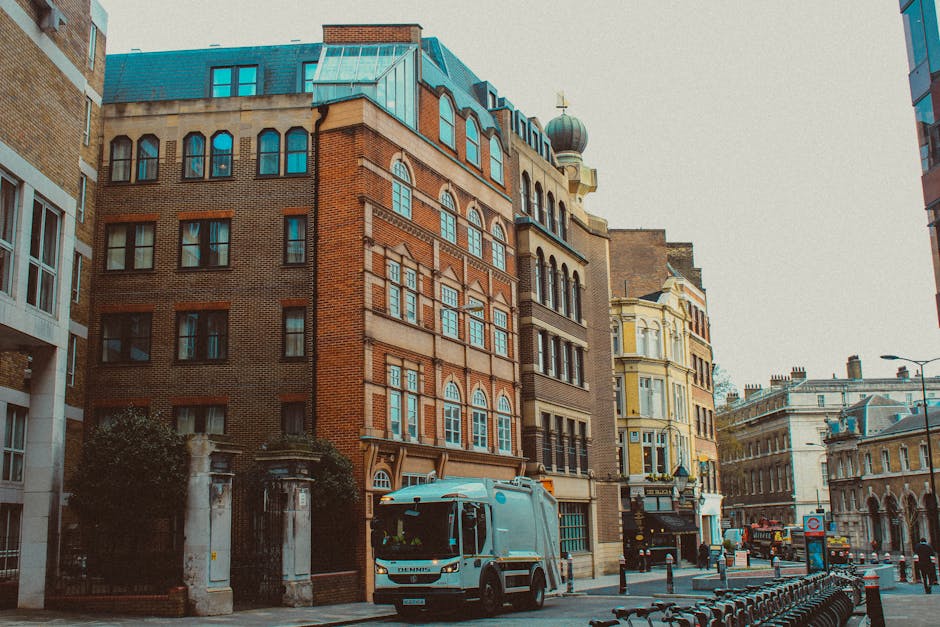 The image depicts a street scene in an urban area with a row of historic multi-storey buildings featuring ornate facades, arched windows, and decorative brickwork in warm tones of red and beige. To the right, a light yellow building with intricate architectural details and large bay windows adds visual interest. A white rubbish collection vehicle, with a sleek design and branding indicating it belongs to a waste disposal service, is parked on the street, positioned towards the middle foreground, ready for waste collection. The vehicle is situated near a sidewalk lined with metal bike racks, and the street surface appears to be made of asphalt. In the background, there are other buildings, street lamps, and a few pedestrians, all under a clear sky with diffused daytime lighting. The scene suggests an alternative to council rubbish collection, aligning with private waste removal services like Rubbish Removal Paddington that handle on-site clearance of bulky waste or construction debris, especially in densely built urban environments where street space and permit rules may limit standard disposal options.