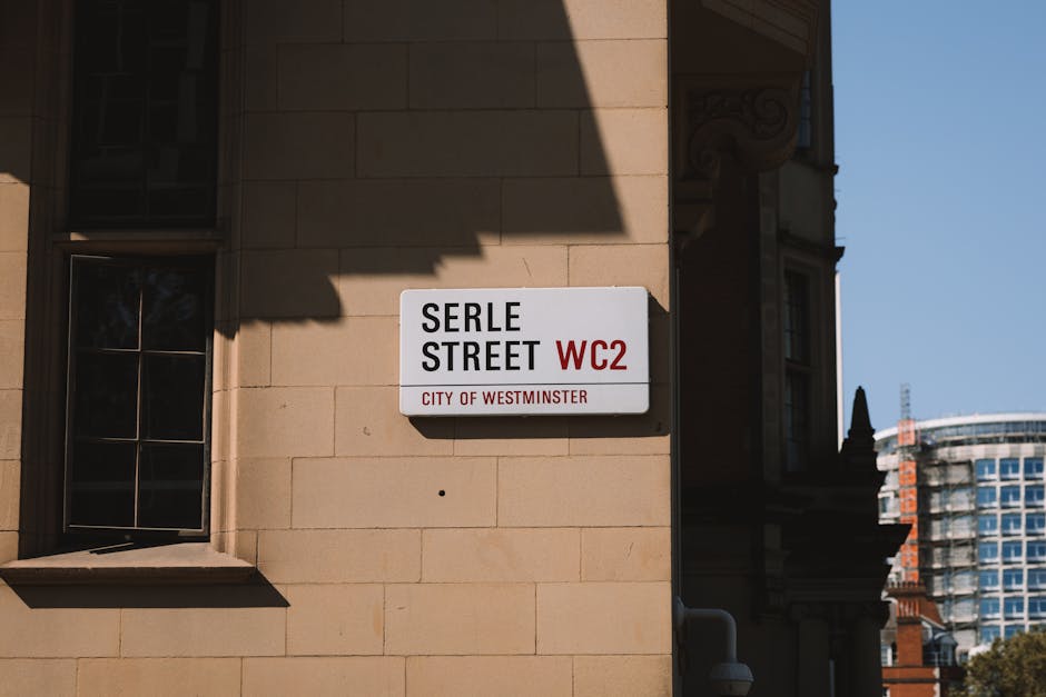 A rectangular street sign mounted on the beige stone exterior wall of a building, indicating 'Serle Street WC2' in black and red text, with 'City of Westminster' written below in smaller red letters. The sign is positioned near a black-framed window with multiple panes, and a shadow from an overhanging structure partially covers the upper part of the wall and sign. In the background, part of an adjacent building with ornate architectural details and a modern high-rise structure with glass windows are visible under a clear blue sky. The scene reflects urban environment characteristics typical of Westminster, with careful attention to the materials and textures of the stone façade and surrounding architecture, subtly aligning with themes of city waste management and alternative rubbish collection services provided by companies like Rubbish Removal Paddington.