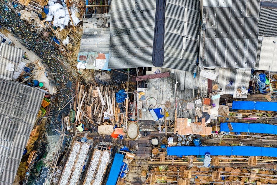 An aerial view of a densely cluttered outdoor area featuring numerous piles of mixed waste and discarded materials. The scene includes a variety of debris such as broken wooden pallets, scattered bricks, and plastic containers. Several sections are covered with rusted corrugated metal sheets, some of which are bent or damaged, forming makeshift roofs or walls. There are also patches of blue tarpaulin used as covers, contrasting with the weathered metal and earthy tones of the wood and soil. The environment appears to be a segregated waste collection or disposal site, with visible pathways and small areas of standing water among the refuse. The overall atmosphere indicates a site where independent rubbish handling or remodeling waste is being managed, aligning with private rubbish removal services such as those offered by Rubbish Removal Paddington. The scene emphasizes a mix of textures, from rough metal surfaces and fractured wood to soft soil and reflective water, captured under natural light that highlights the disorderly arrangement of waste materials.