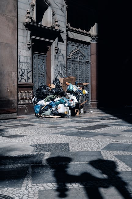 A street scene featuring a red rubbish collection vehicle with its rear compactor open, positioned on the roadside. A worker wearing a high-visibility yellow and red vest is standing behind the vehicle, handling black rubbish bags placed on a small, wheeled trolley. The waste bags appear full and are being prepared for disposal. The vehicle is situated on a paved urban road with visible lane markings, and nearby on the sidewalk, there is a black motorcycle parked next to the trolley. In the background, trees and utility poles are visible, with overcast skies creating a diffused lighting environment. The surrounding environment suggests an area where independent waste collection or alternative rubbish disposal services, like those provided by Rubbish Removal Paddington, are utilized for house or commercial clearance. The scene depicts routine waste management activity in a suburban or urban setting, emphasizing the handling of domestic or commercial rubbish before collection or transfer to disposal sites.
